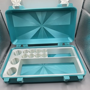 May include: A rectangular turquoise storage case with a hinged lid featuring a sunburst pattern. The interior contains a white insert with multiple circular compartments of varying sizes and two rectangular sections for organized storage.