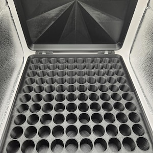 May include: A black, square storage case with an open lid featuring a geometric starburst pattern. The interior base contains a grid of one hundred circular compartments arranged in a ten-by-ten pattern, designed for holding small, cylindrical items.