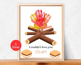 May include: Framed print featuring a campfire design made with handprints in red, orange, and yellow. The campfire sits above brown logs and the text "I couldn't love you s'more" with two s'mores below.