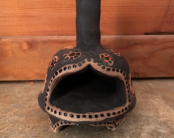 Pode incluir: Um pequeno pote de cerâmica preto com uma chaminé, decorado com pontos laranja e castanho.