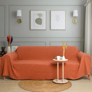 May include: A burnt orange sofa cover with a textured waffle-knit pattern draped over a couch. The cover features a consistent grid-like weave across the entire surface, providing a uniform appearance that fits snugly over the cushions and arms.