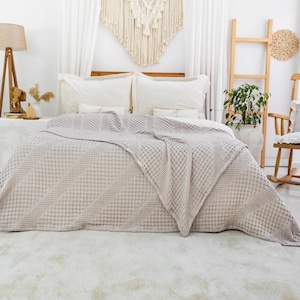 May include: A light beige waffle-weave bedspread draped over a bed with two white pillows. The fabric features a textured grid pattern throughout, covering the mattress and extending down to the floor in a soft, folded drape across the surface.