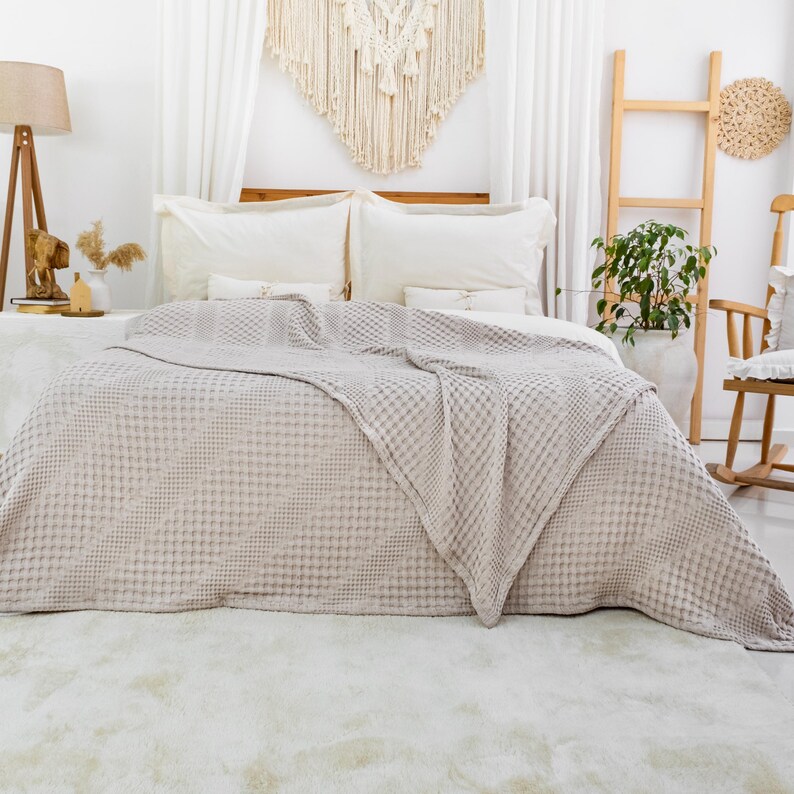 May include: A light beige waffle-weave bedspread draped over a bed with two white pillows. The fabric features a textured grid pattern throughout, covering the mattress and extending down to the floor in a soft, folded drape across the surface.
