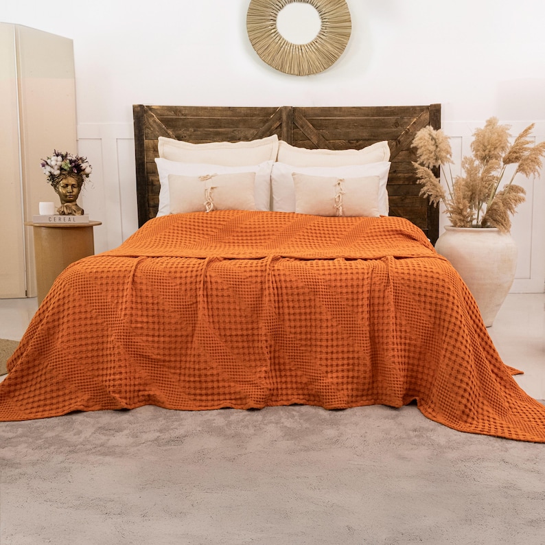 May include: A vibrant orange waffle-weave bedspread draped over a bed with a dark brown wooden headboard. The bed features two large white pillows and two smaller rectangular accent pillows with tassels, creating a textured bedding arrangement.