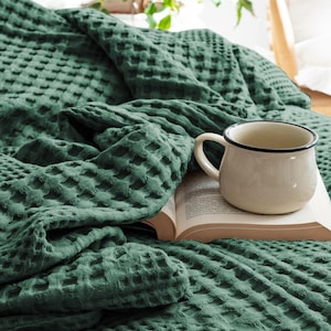 May include: A dark green waffle-weave blanket draped in folds next to an open book and a cream-coloured ceramic mug with a dark rim. The textured fabric features a repeating square grid pattern, creating a soft and layered appearance across the surface.