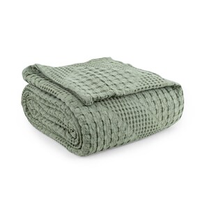 May include: A sage green blanket featuring a distinct waffle weave texture. The fabric is folded into a neat, rectangular stack, showcasing the uniform grid pattern across the entire surface of the textile. The material has a soft, matte appearance.
