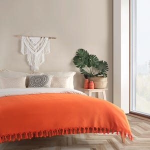 Puede incluir: Una colcha de color naranja brillante con un borde de flecos en la parte inferior, que cubre una cama con almohadas blancas y un coj&iacute;n decorativo con patr&oacute;n circular. Un tapiz de macram&eacute; blanco cuelga sobre la cama junto a una planta verde.
