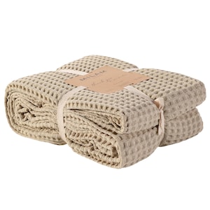 May include: A folded beige waffle-weave blanket secured with a light-coloured ribbon and a rectangular paper tag. The tag features the text MILAM and the phrase thank you for your purchase printed in black ink on the front surface of the paper.