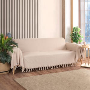 May include: A cream-coloured sofa with a fringe trim throw blanket. The sofa is in a living room with a striped wall and a rug on the floor.