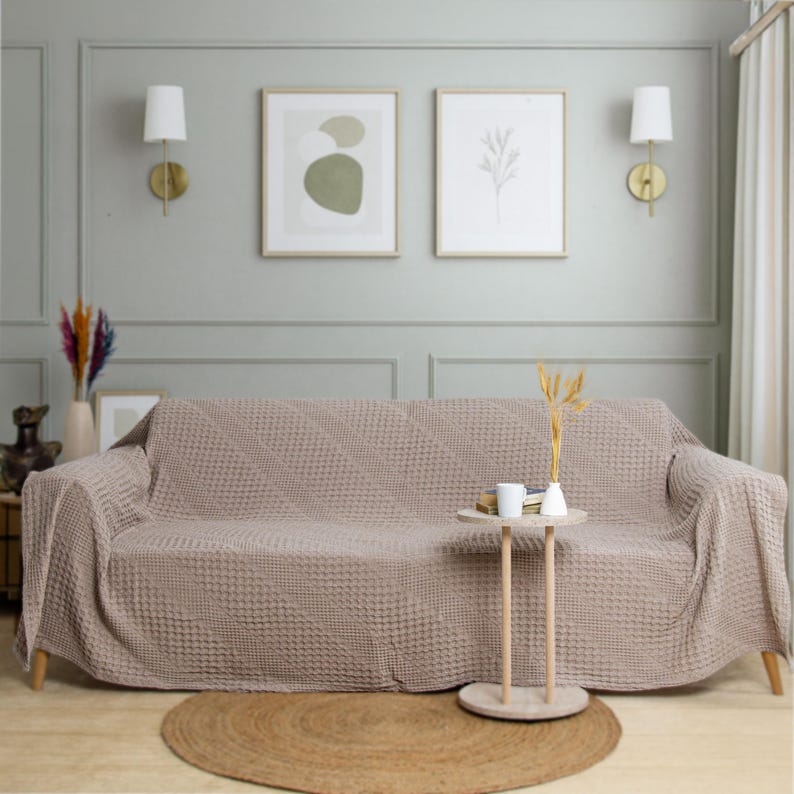 May include: A light taupe sofa cover with a textured waffle-knit pattern draped over a three-seater couch. The fabric features a consistent grid-like weave throughout, providing a uniform appearance across the backrest, seat, and armrests.