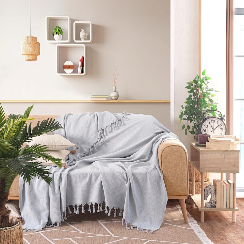 Puede incluir: Una manta tejida de color gris claro con borlas decorativas en los bordes, colocada sobre un sof&aacute; de color beige. La manta presenta un sutil patr&oacute;n texturizado y cubre los cojines del asiento y el respaldo del sof&aacute;, aportando un toque neutro.