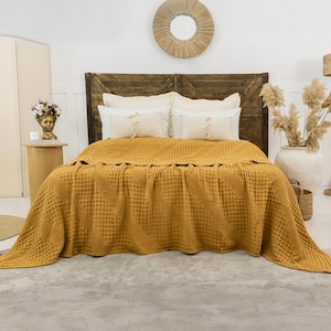 May include: A mustard yellow waffle-weave bedspread draped over a bed with a dark brown wooden headboard. The bed features two large white pillows and two smaller rectangular accent pillows with fringe details centered on the bedspread.
