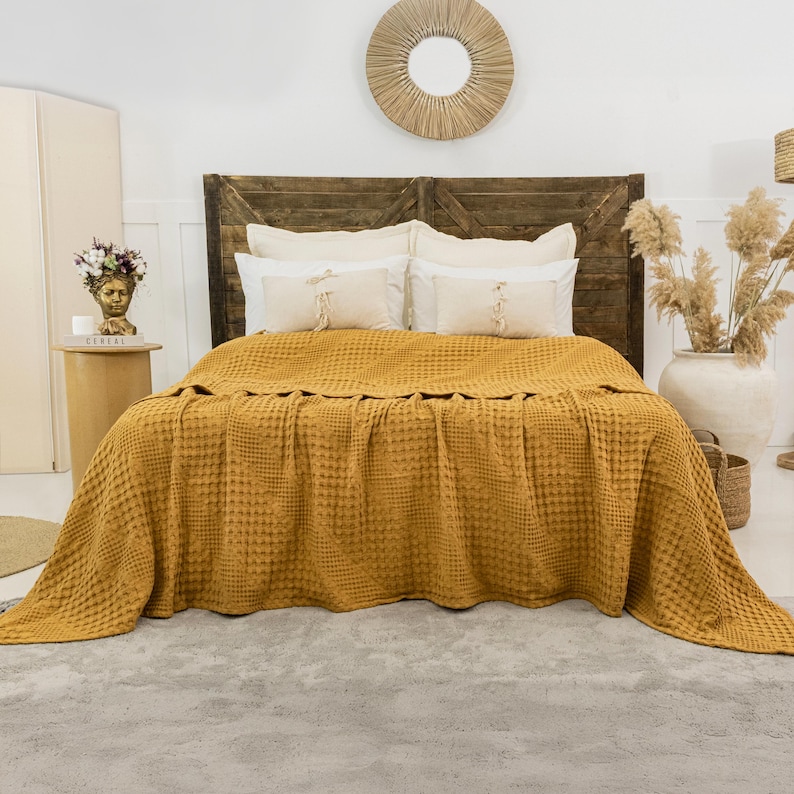 May include: A mustard yellow waffle-weave bedspread draped over a bed with a dark brown wooden headboard. The bed features two large white pillows and two smaller rectangular accent pillows with fringe details centered on the bedspread.