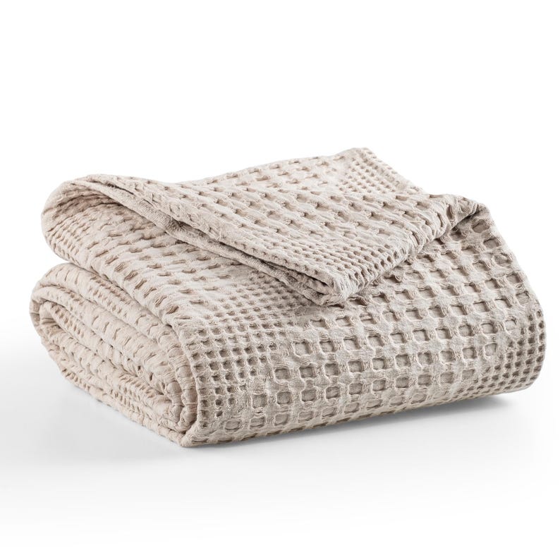 May include: A folded beige blanket featuring a distinct waffle weave texture. The fabric has a grid-like pattern created by the raised square design throughout the material. The blanket is neatly stacked, showing multiple layers of the textured textile.