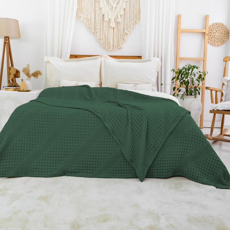 May include: A dark forest green blanket with a textured waffle weave pattern draped over a bed. The blanket features a consistent grid-like surface structure and covers the majority of the bed frame, extending down to the floor in a soft, folded drape.