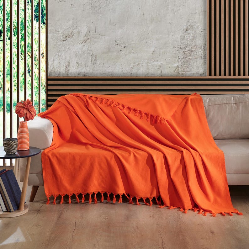 Puede incluir: Una manta de color naranja vibrante colocada sobre un sof&aacute; de tono claro. La manta presenta una textura suave y est&aacute; rematada con borlas decorativas a lo largo del borde inferior y una fila de borlas en la parte central de la tela.