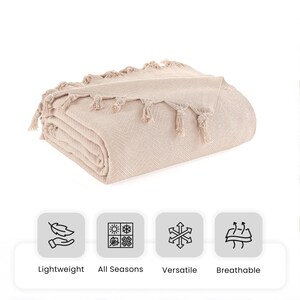 Puede incluir: Una manta beige doblada con un patr&oacute;n de tejido en espiga y borlas decorativas en los bordes. Debajo de la manta hay cuatro iconos etiquetados como Lightweight, All Seasons, Versatile y Breathable, que representan las caracter&iacute;sticas.