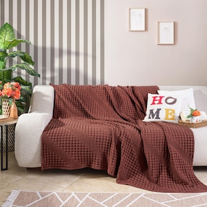 May include: A textured, deep rust-coloured waffle-weave throw blanket draped over a light-coloured sofa. A white square throw pillow with the word HOME printed in red and yellow letters sits on the sofa next to the blanket, adding a decorative touch.