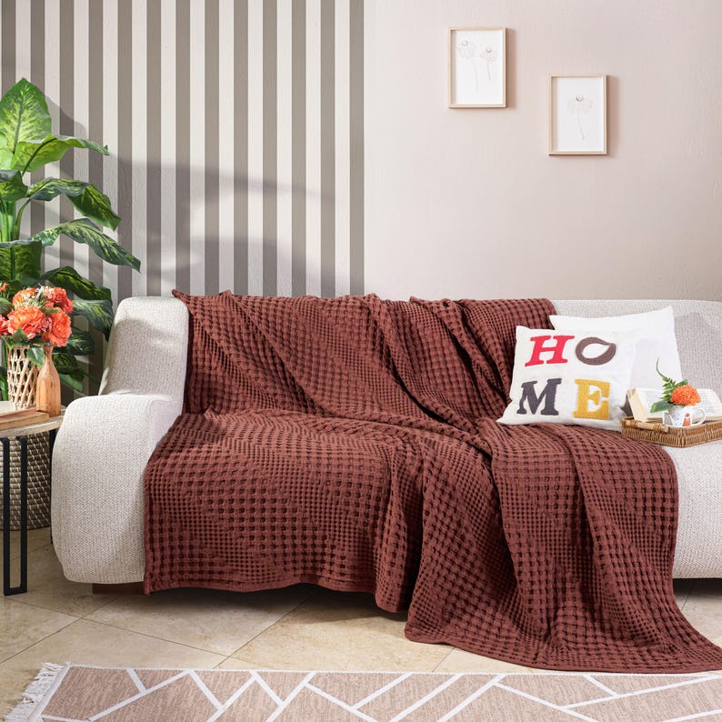 May include: A textured, deep rust-coloured waffle-weave throw blanket draped over a light-coloured sofa. A white square throw pillow with the word HOME printed in red and yellow letters sits on the sofa next to the blanket, adding a decorative touch.