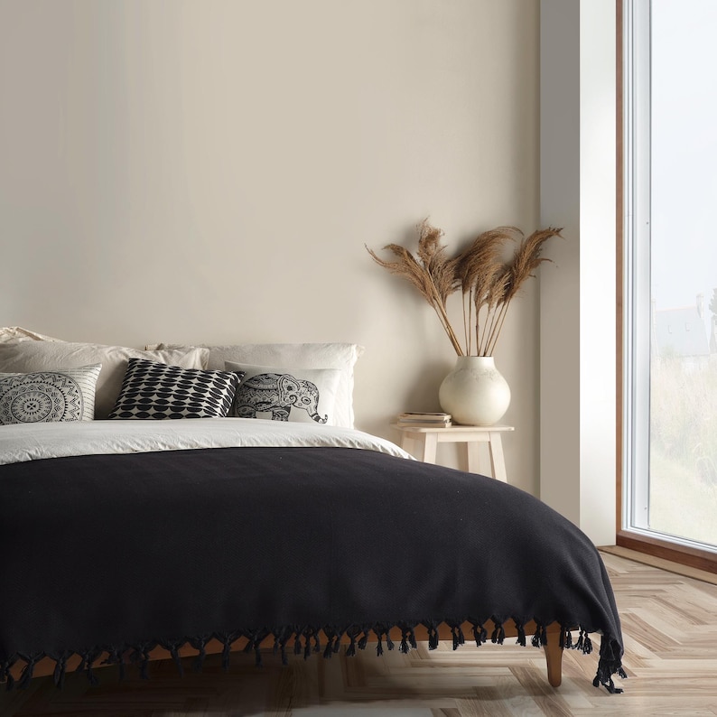 Puede incluir: Una manta negra lisa con bordes de flecos anudados colocada sobre una cama. La cama incluye almohadas blancas, una almohada con patr&oacute;n circular en blanco y negro, una almohada geom&eacute;trica en blanco y negro y una almohada con un elefante.
