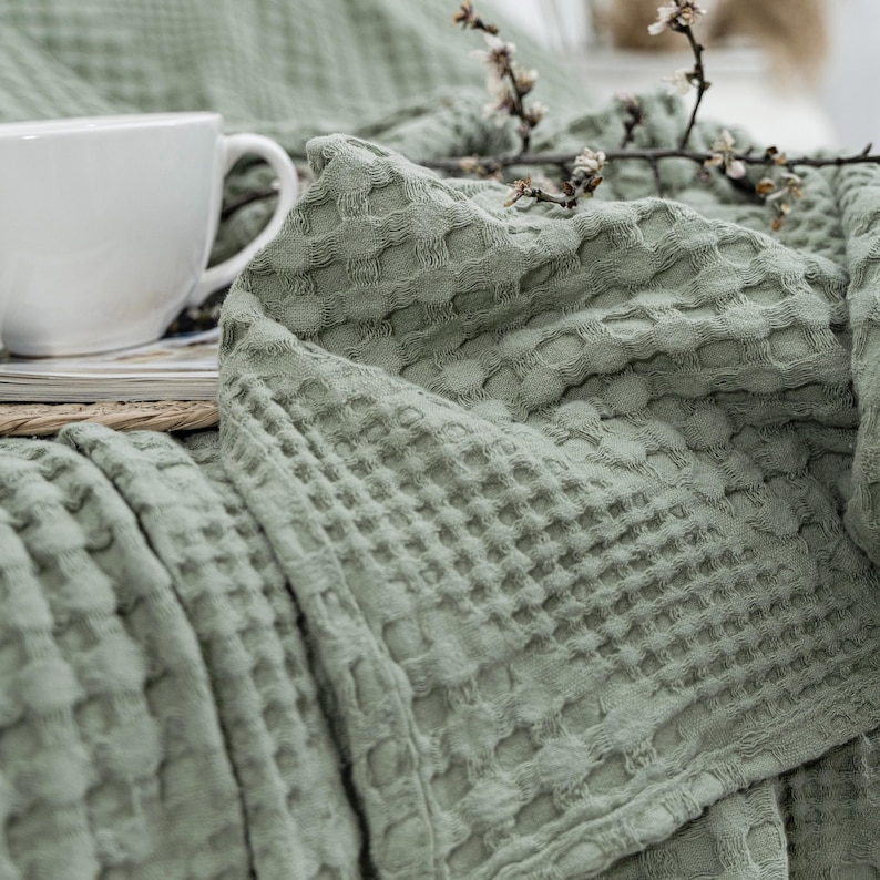May include: A sage green waffle-weave blanket with a textured grid pattern. A white ceramic mug sits on a stack of books partially underneath the blanket, and a small branch with white blossoms rests on top of the folded fabric folds.