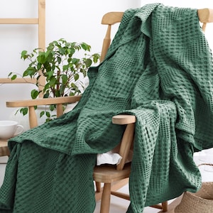 May include: A deep forest green blanket featuring a textured waffle weave pattern. The blanket is draped over a light-coloured wooden chair, showcasing its square grid design and soft, folded edges that cascade down the seat and back of the furniture.