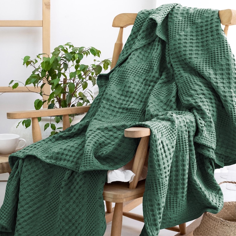 May include: A deep forest green blanket featuring a textured waffle weave pattern. The blanket is draped over a light-coloured wooden chair, showcasing its square grid design and soft, folded edges that cascade down the seat and back of the furniture.