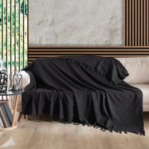 Puede incluir: Una manta de color negro s&oacute;lido colocada sobre un sof&aacute; de tono claro. El tejido rectangular presenta una textura suave y tejida, rematada con flecos anudados decorativos a lo largo de los bordes, creando un aspecto sencillo y uniforme.