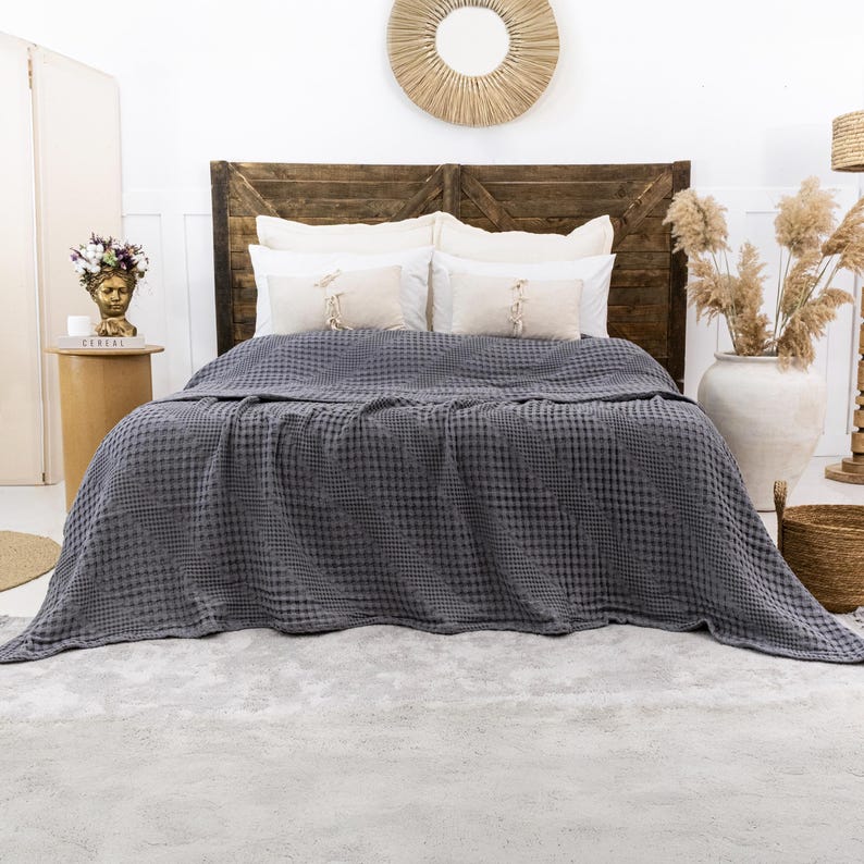 May include: A dark gray waffle-weave blanket draped over a bed. The blanket features a textured grid pattern across its surface. The bed is styled with two large white pillows and two smaller rectangular accent pillows with central ties.