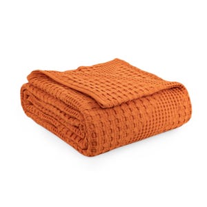 May include: A folded blanket in a solid burnt orange colour featuring a distinct waffle weave texture. The fabric has a grid-like pattern across the entire surface, with a finished hem along the top edge, creating a structured and uniform appearance.