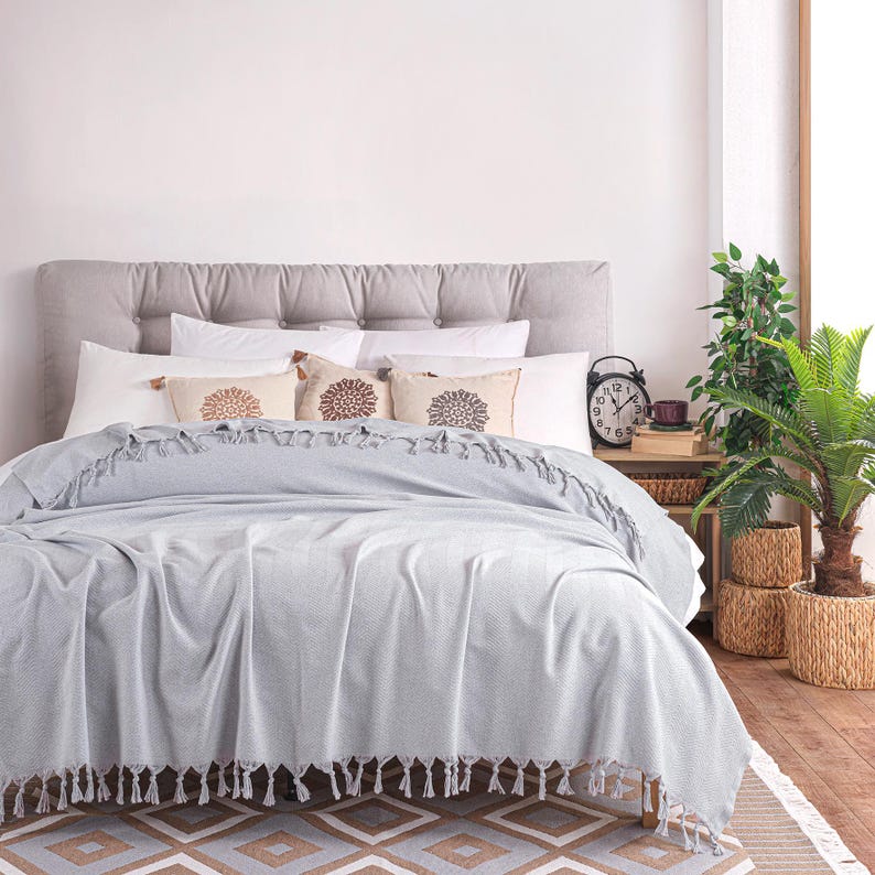 Puede incluir: Una manta de color gris claro con textura y flecos colocada sobre una cama. La cama cuenta con un cabecero gris capiton&eacute;, almohadas blancas y tres cojines beige peque&ntilde;os con patrones circulares marrones. Un despertador negro est&aacute; al lado.