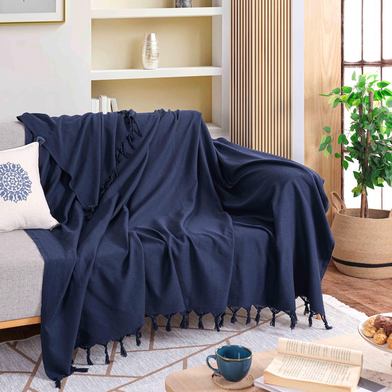 Puede incluir: Una manta de color azul marino s&oacute;lido con un tejido texturizado y flecos anudados decorativos en los bordes, colocada sobre un sof&aacute; gris claro. Un coj&iacute;n blanco con un patr&oacute;n circular azul estilo mandala se sit&uacute;a en el lado izquierdo.