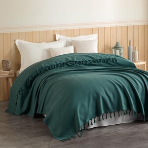 Puede incluir: Una colcha de color verde azulado oscuro con un tejido texturizado y flecos en los bordes, colocada sobre una cama con almohadas blancas y de color blanco roto. La colcha presenta un patr&oacute;n uniforme y cuelga por los lados de la cama.
