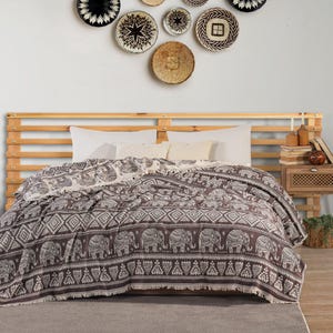 May include: A dark brown and white woven throw blanket featuring a repeating pattern of elephants and geometric shapes with a fringed edge, draped over a bed with white pillows and a light wood headboard against a wall with decorative baskets.