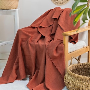 May include: A rust-coloured throw blanket with a subtle herringbone weave pattern and frayed edges draped over a wooden chair. The fabric has a textured appearance and covers the seat and backrest of the chair, showing a consistent deep orange tone.