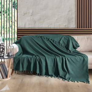 May include: A deep green throw blanket with fringe edges draped over a white sofa. The blanket is made of a soft, textured fabric.