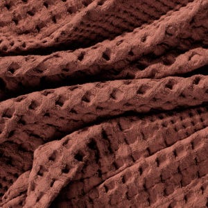 May include: A terracotta-coloured fabric featuring a distinct waffle weave texture. The material is folded, creating deep shadows and highlights that emphasise the grid-like pattern of the textile surface across the entire frame of the image.