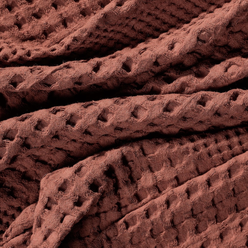 May include: A terracotta-coloured fabric featuring a distinct waffle weave texture. The material is folded, creating deep shadows and highlights that emphasise the grid-like pattern of the textile surface across the entire frame of the image.