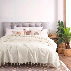Puede incluir: Una colcha de color crema con un borde de flecos, colocada sobre una cama con un cabecero gris capiton&eacute;. Tres cojines rectangulares decorativos con bordados circulares marrones estilo mandala est&aacute;n dispuestos contra almohadas blancas.