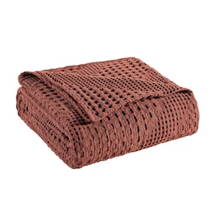 May include: A folded blanket in a deep terracotta red color featuring a prominent waffle weave texture. The fabric has a grid-like pattern created by the raised square sections of the weave, giving the textile a thick and dimensional appearance.