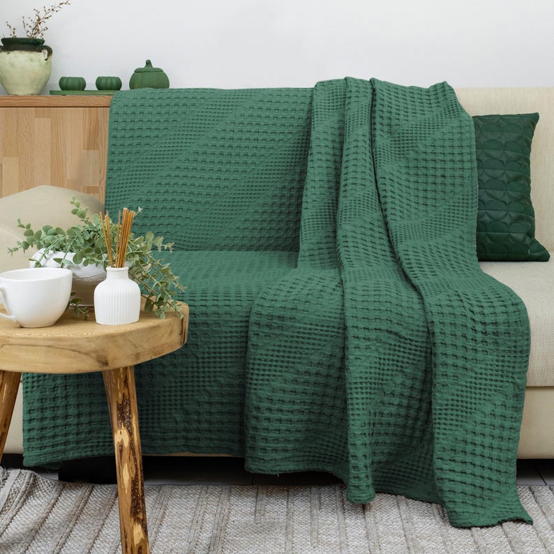 May include: A dark green waffle-weave throw blanket draped over a sofa. The blanket features a distinct grid-like texture throughout its surface. A matching dark green throw pillow is positioned on the right side of the sofa, completing the set.