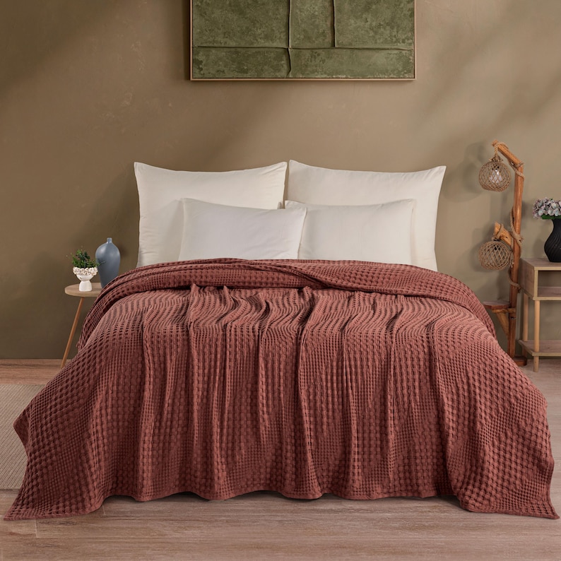 May include: A terracotta-colored waffle weave bedspread draped over a bed with two large white pillows and two smaller white pillows. The textured fabric features a grid-like pattern, covering the mattress and extending down to the floor.