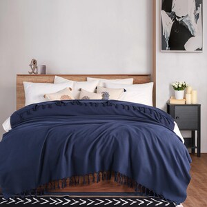 Puede incluir: Una manta de color azul marino s&oacute;lido con borlas decorativas en los bordes superior e inferior. La manta est&aacute; colocada sobre una cama con almohadas blancas y cojines m&aacute;s peque&ntilde;os con patrones circulares, frente a un cabecero de madera.