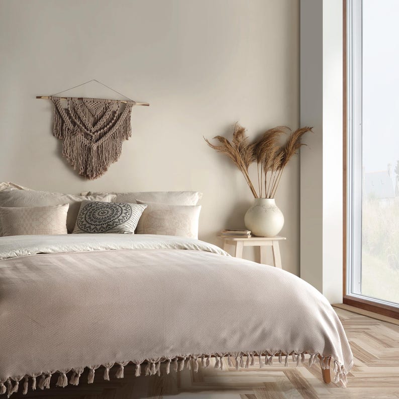 Puede incluir: Una colcha beige con un patr&oacute;n texturizado y borde de flecos, sobre una cama con varias almohadas, incluida una con un dise&ntilde;o de mandala circular en blanco y negro. Tambi&eacute;n hay un tapiz de macram&eacute; y un jarr&oacute;n con hierbas secas.