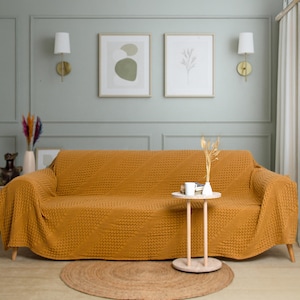 May include: A mustard yellow sofa cover with a textured waffle-knit pattern draped over a couch. The fabric features a consistent grid-like design across the entire surface, providing a uniform look that covers the back, seat, and armrests.