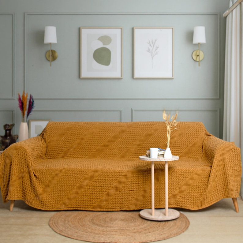May include: A mustard yellow sofa cover with a textured waffle-knit pattern draped over a couch. The fabric features a consistent grid-like design across the entire surface, providing a uniform look that covers the back, seat, and armrests.