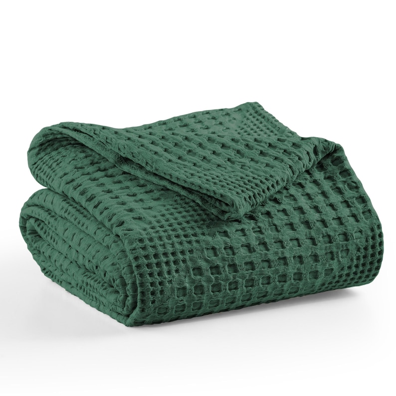 May include: A dark forest green blanket featuring a prominent waffle weave texture. The fabric is folded into a neat, layered stack, showcasing the square grid pattern across the entire surface of the textile. The edges are finished with a hem.
