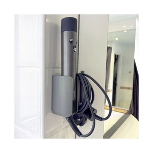 Puede incluir: Un soporte de pared gris con un secador de pelo Dyson gris y un cable negro. El soporte tiene la palabra "Dyson" impresa.