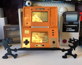 Puede incluir: Una consola de juegos portátil Nintendo Game & Watch vintage de color naranja y dorado con el juego Donkey Kong mostrado en la pantalla. La consola está en un soporte negro con dos figuras negras a cada lado.