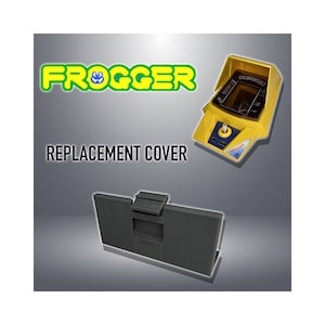 Puede incluir: Una cubierta de repuesto para un juego de Frogger vintage amarillo de mano. La cubierta es negra y tiene un pequeño pestillo con la palabra "ABRIR" impresa en ella.